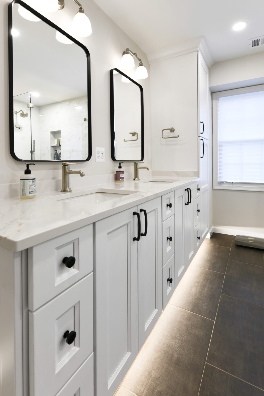 Vienna Primary En-suite and Laundry (7) Modern bathroom with dual sinks, white cabinetry, black hardware, rectangular mirrors, wall-mounted lights, and under-cabinet lighting on a dark tile floor.