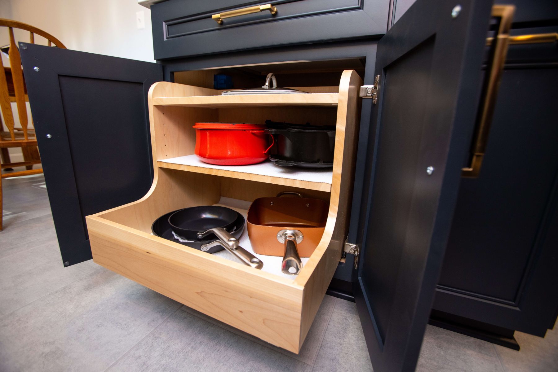 Open kitchen cabinet with a wooden pull-out shelf holding cookware, including pans, a red pot, a black pot, and a copper-colored pot.