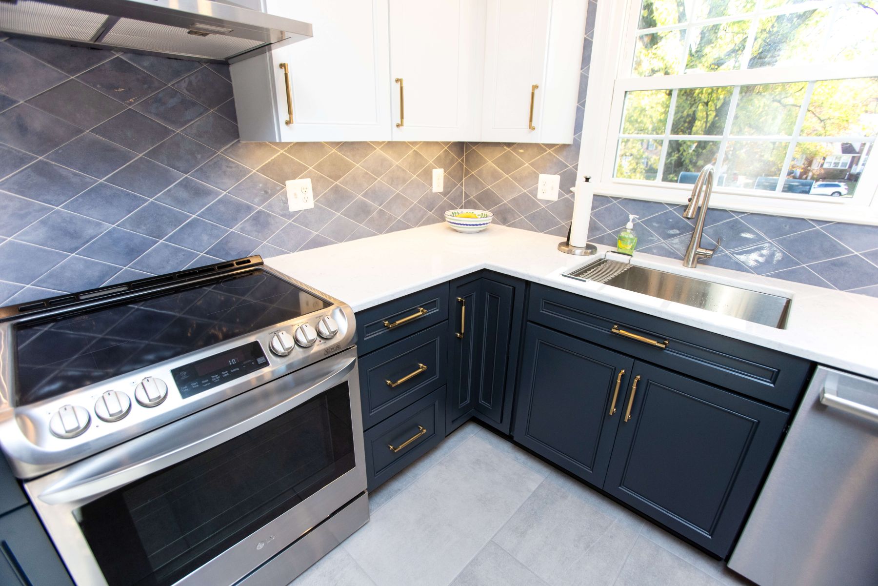 Modern kitchen with stainless steel stove, dark blue lower cabinets, white upper cabinets, large sink, and a blue tiled backsplash near a window.