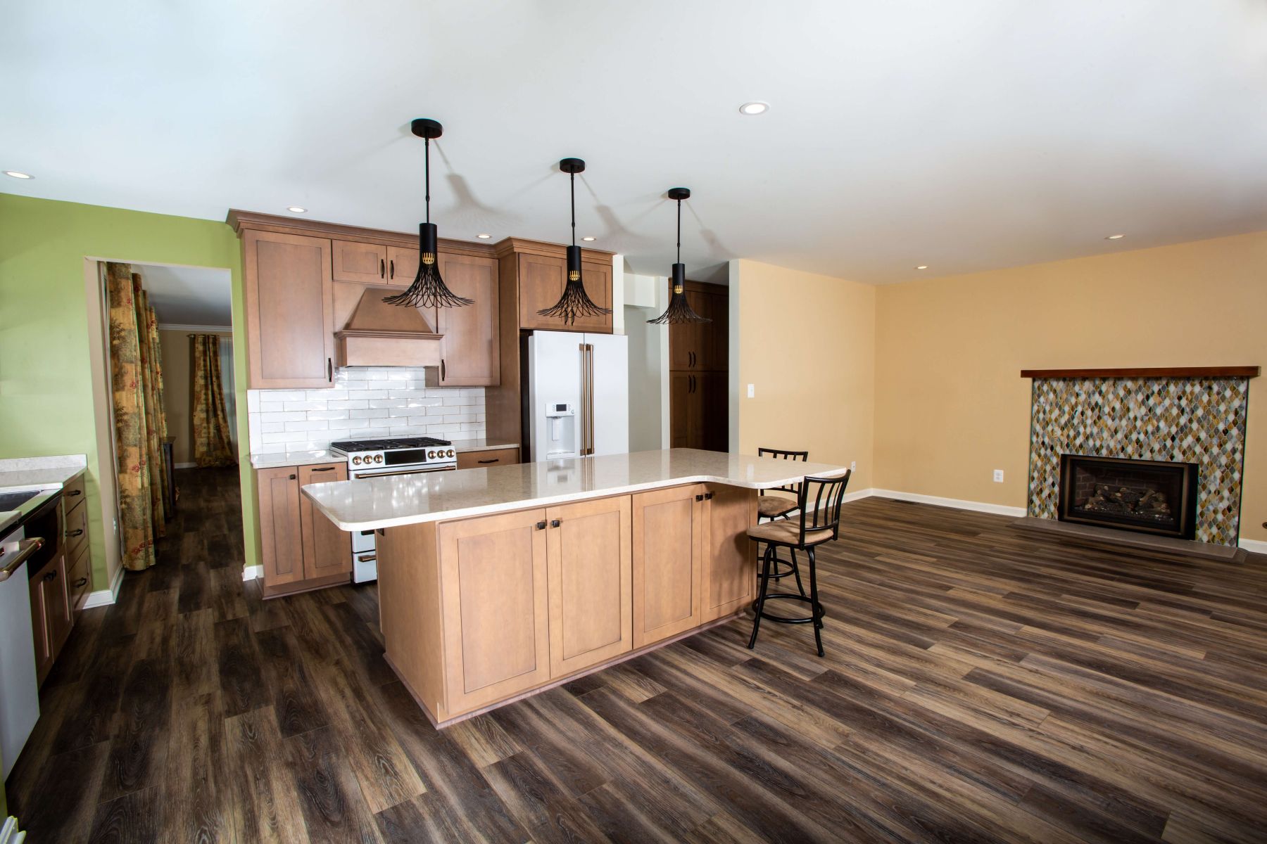 Modern kitchen with wood cabinets, white countertops, an island with stools, stainless steel appliances, and an open living area with a fireplace and wood-style flooring.