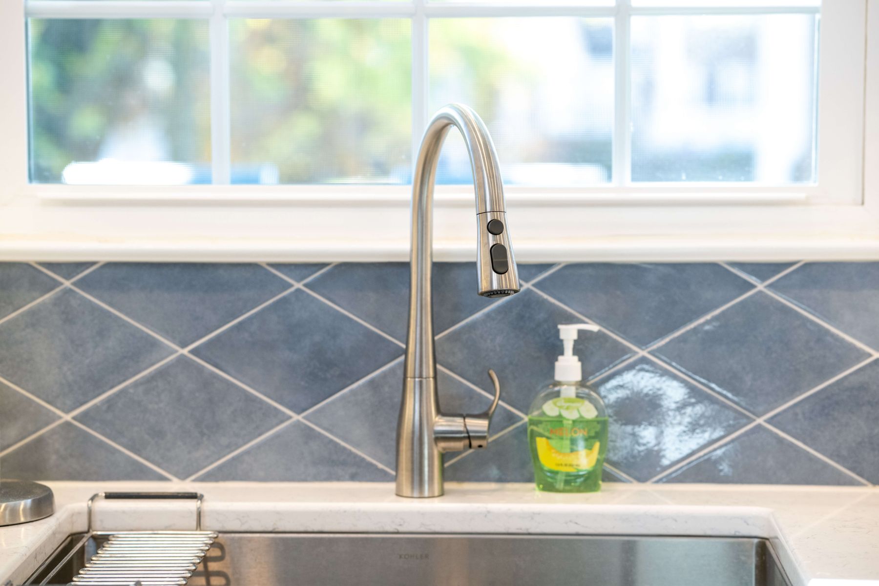 A stainless steel kitchen faucet with a soap dispenser sits on a white countertop in front of a blue tiled backsplash and a window.