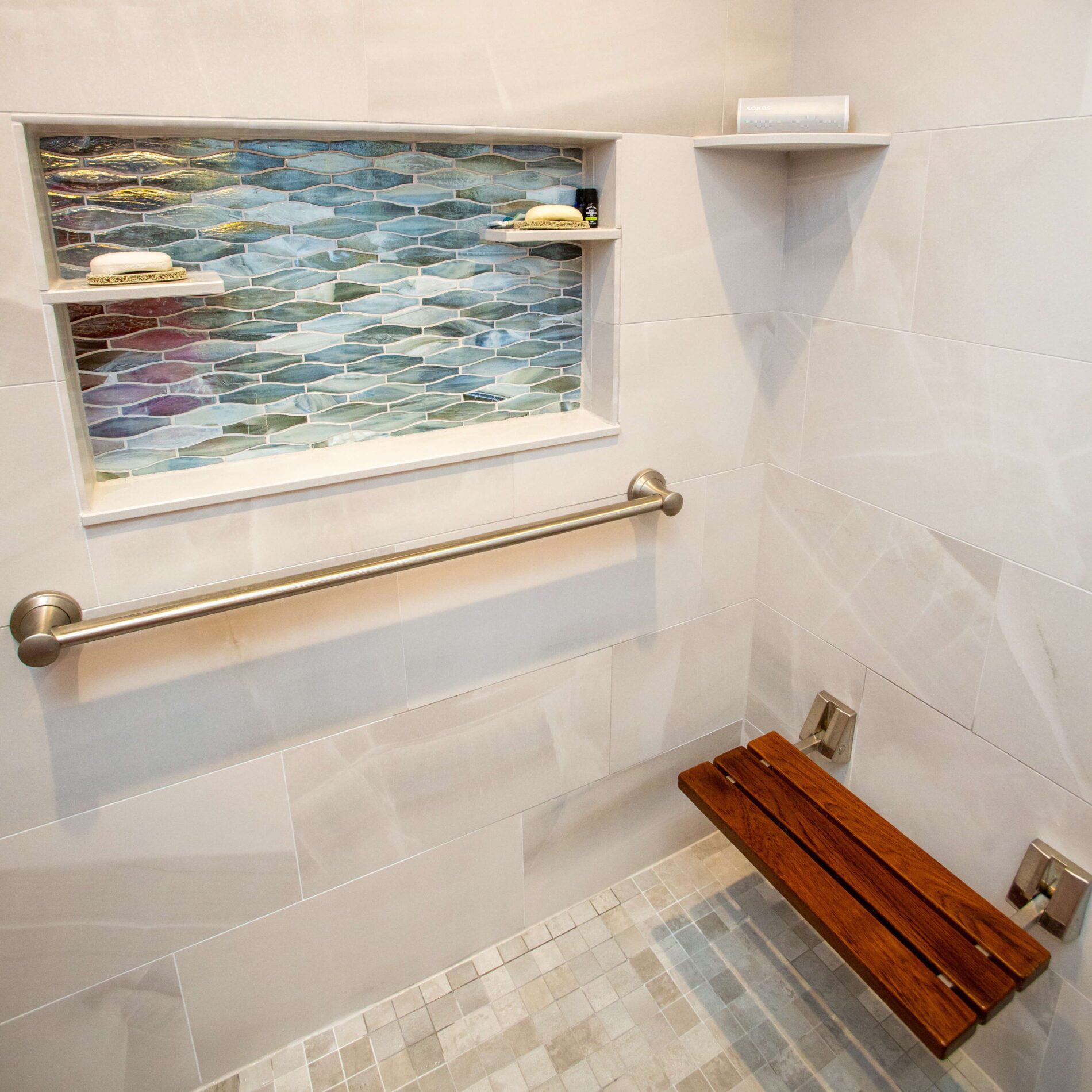 Shower with a wall-mounted grab bar, a foldable wooden bench, a niche with blue tile accents, and a corner shelf holding soap and toiletries—ideal aging in place products for comfort and safety.