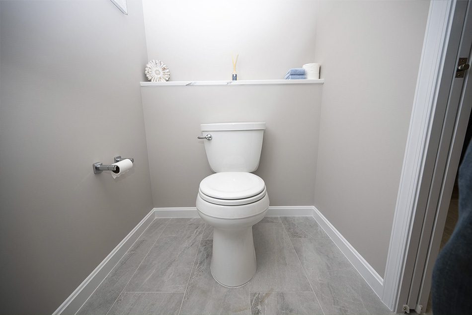 A modern white toilet is centered in a small bathroom with light gray tiled floor and beige walls—perfect for aging in place remodeling in Northern Virginia. A shelf behind the toilet holds decor, a towel, and toilet paper.