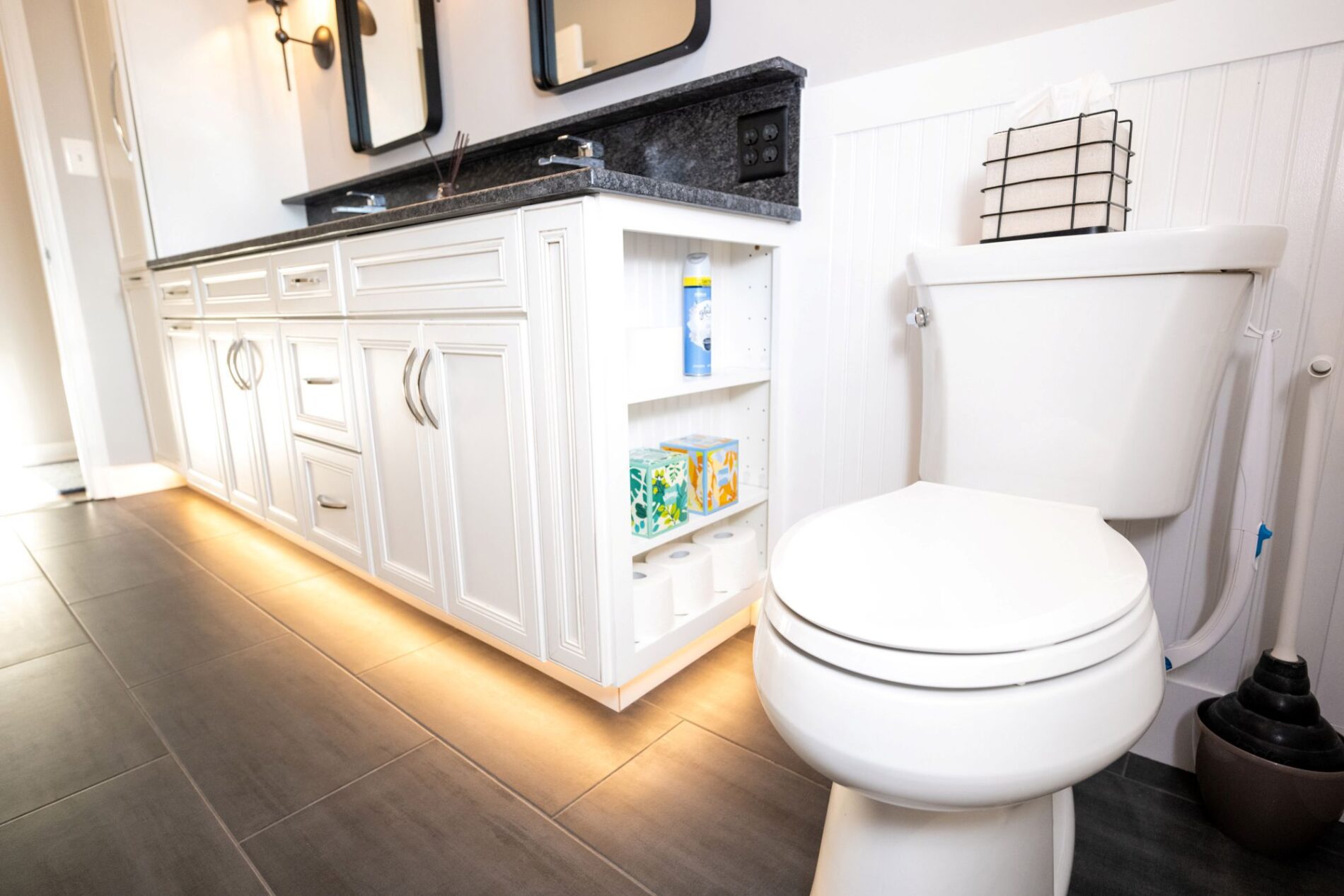 A modern bathroom with a white toilet, built-in vanity, aging in place products, open shelving with tissues and toilet paper, and under-cabinet lighting on a gray tiled floor.