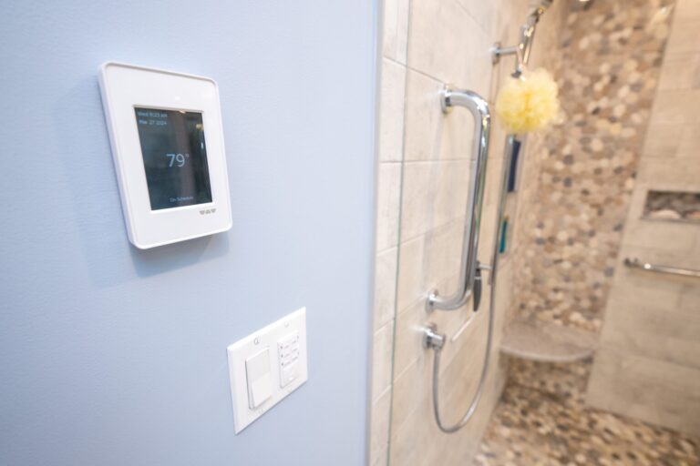 A bathroom with a digital wall thermostat set to 79°F, light switches below, and a shower featuring grab bars and a tiled bench in the background—showcasing the benefits of aging in place.