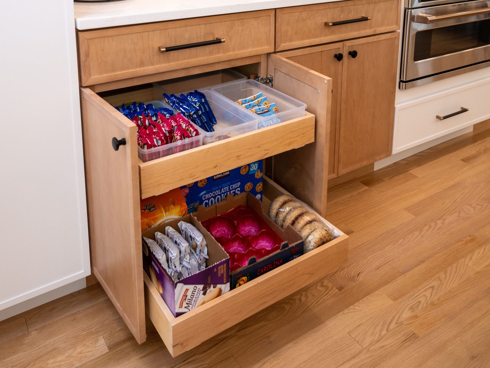 A kitchen cabinet with two pull-out shelves, organized with snacks including cookies, crackers, chips, and bagels in bins and boxes.