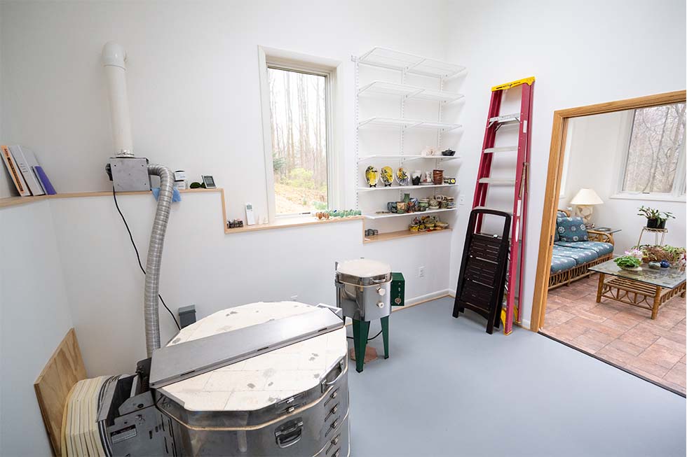 A small studio room with two pottery kilns, wall shelves holding supplies, a red ladder, and a window showing trees outside; an adjoining sunroom is visible through an open door.