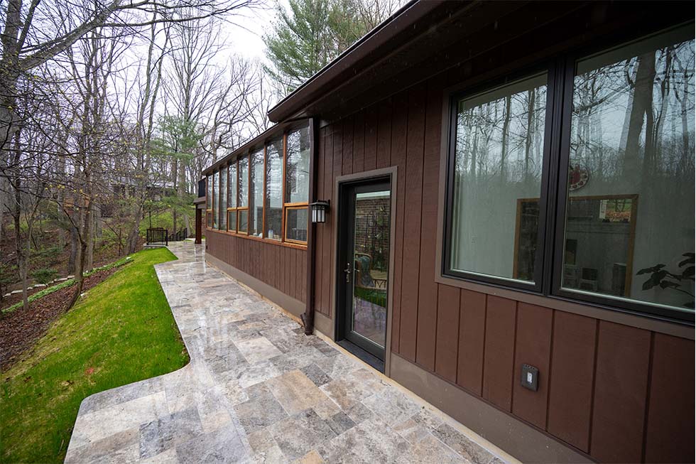 A modern brown house with large windows and a glass door, bordered by a stone walkway and surrounded by trees and grass.