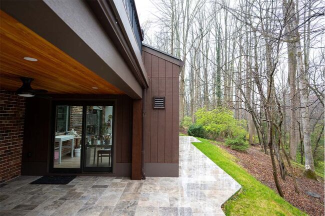 A covered patio with stone tile flooring leads to a walkway beside a brown house, bordered by green grass and trees in a wooded area.