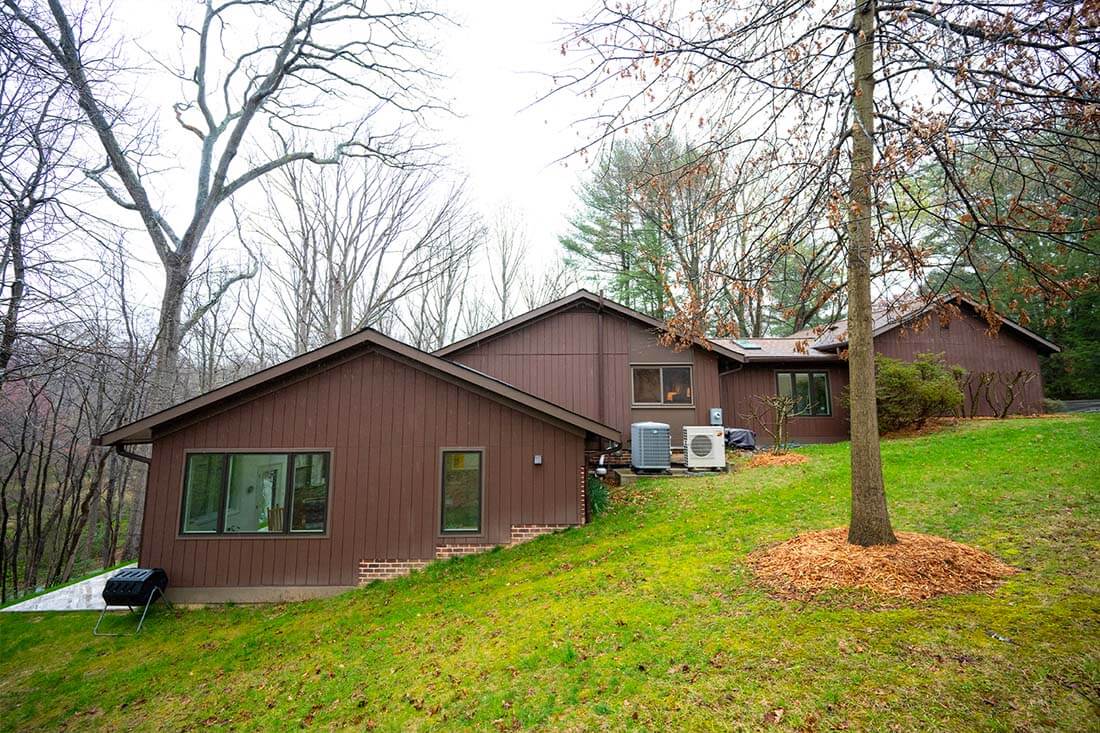 A charming brown house with large windows graces its surroundings of lush trees and grass, featuring two exterior HVAC units on the side, exemplifying the elegant home additions often seen in Reston, Virginia.