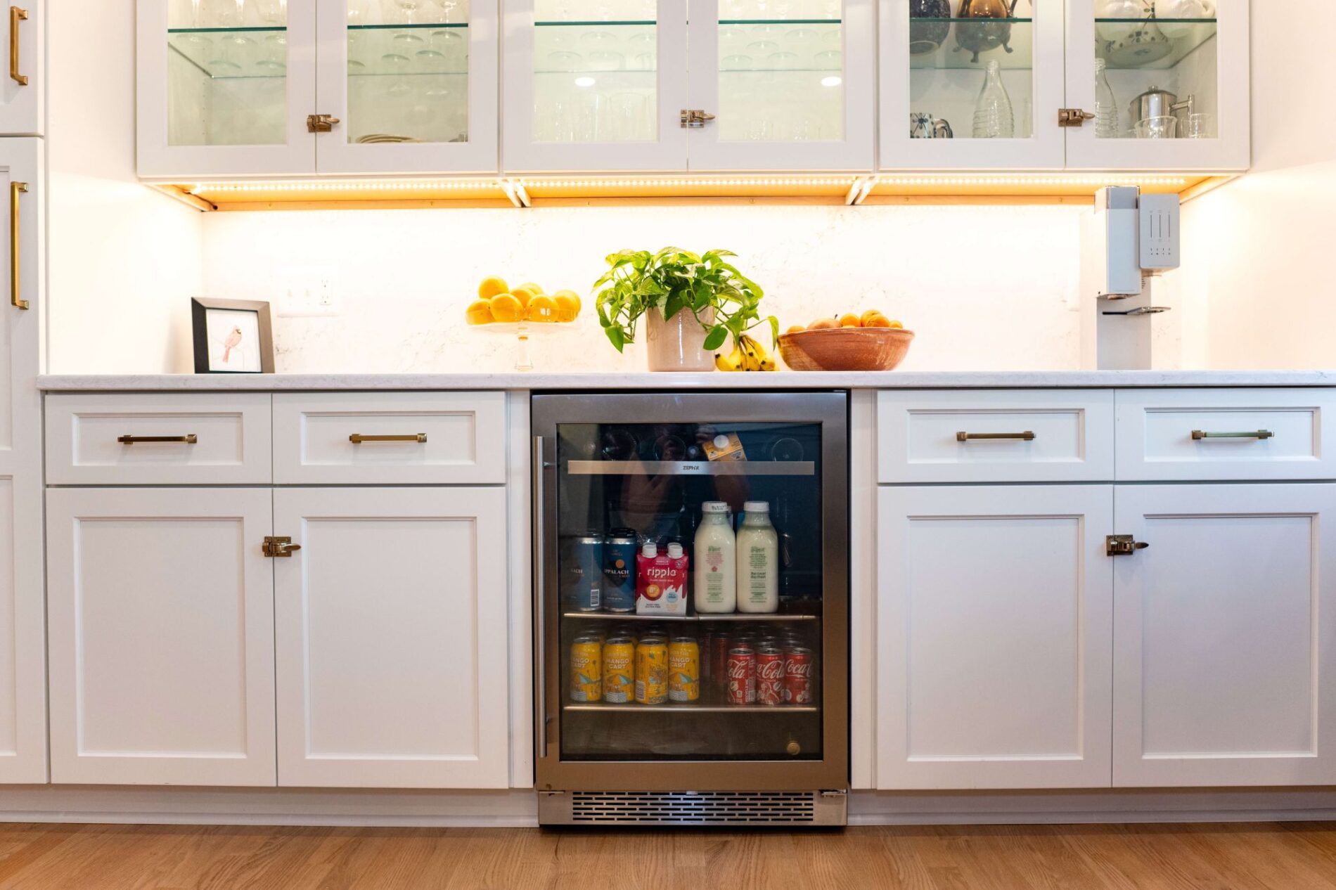 A modern white kitchen in a Northern VA home features a glass-front mini fridge stocked with drinks and milk, fruit on the counter, and cabinets with brass handles—perfect inspiration for your next remodeling project.