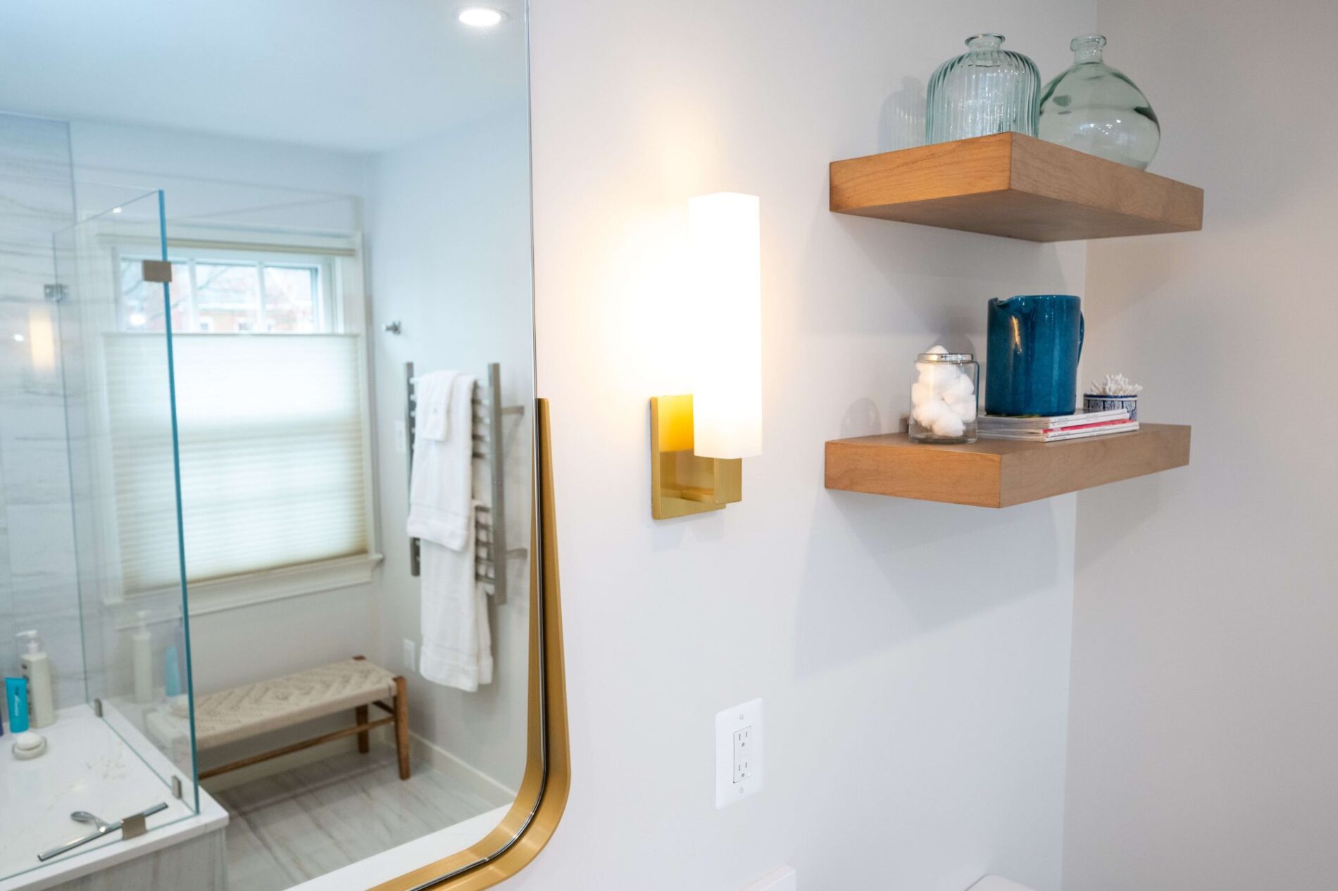 Modern bathroom with wooden floating shelves holding decorative glass vases, a blue jar, cotton balls, and books—perfect inspiration for your Northern VA remodeling project; a light fixture and part of a mirror are also visible.