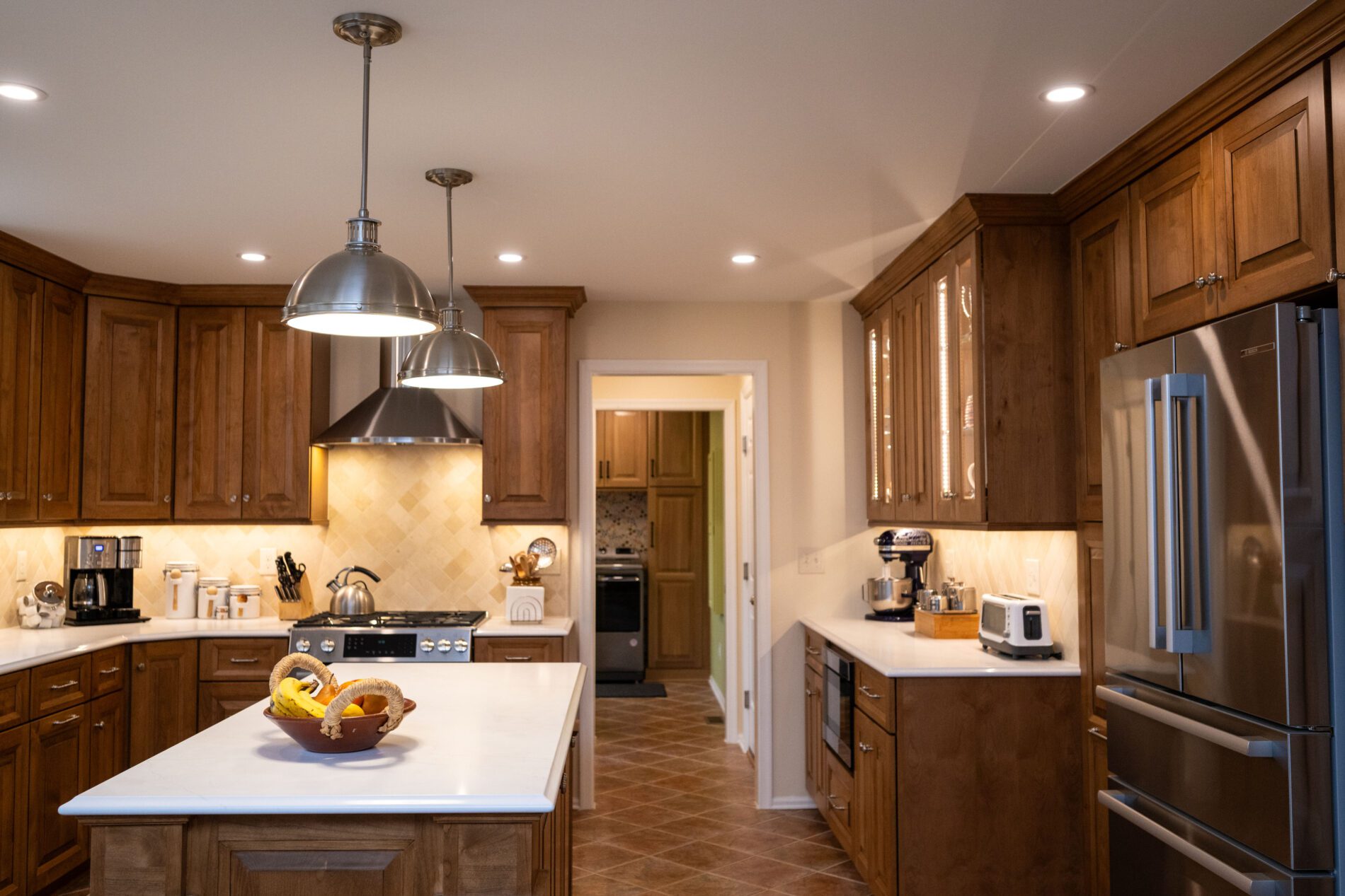 A modern kitchen with wooden cabinets, stainless steel appliances, two hanging lights, and a fruit bowl on the central island invites you to consider: remodel now or wait, especially with a laundry room visible through the back doorway.