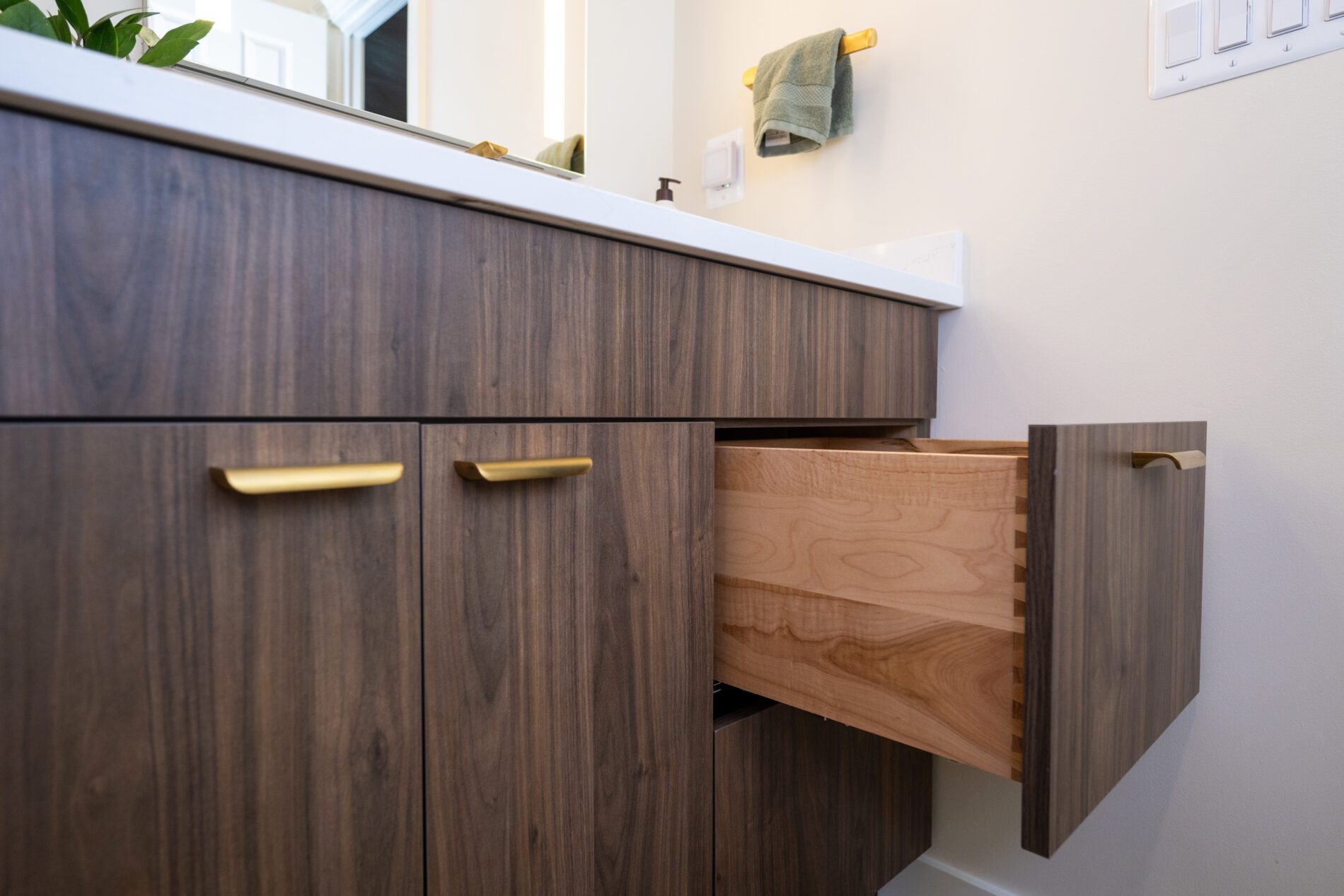 Wood bathroom vanity with gold handles, one drawer partially open showing dovetail joints, light countertop, and a green towel hanging on the wall—perfect for those remodeling Northern Virginia despite rising costs.