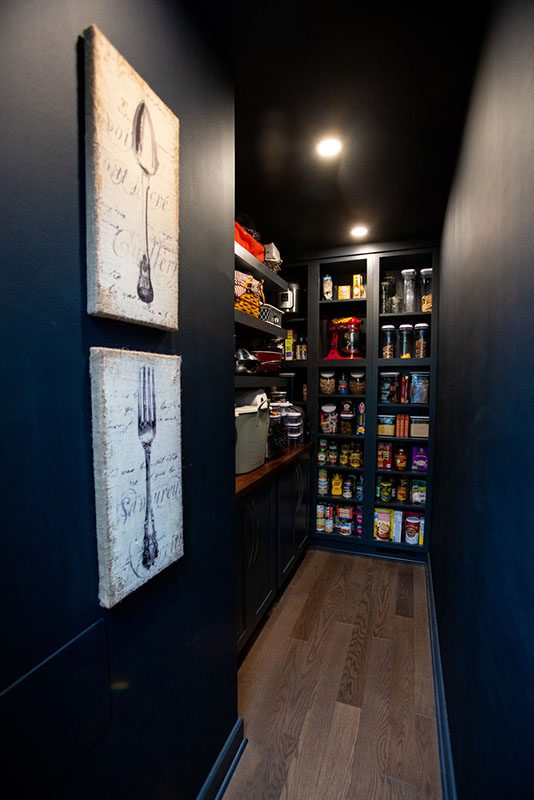 A narrow pantry with dark walls and wooden floor, featuring shelves stocked with canned goods, jars, and kitchen supplies; two utensil-themed artworks hang on the left wall.