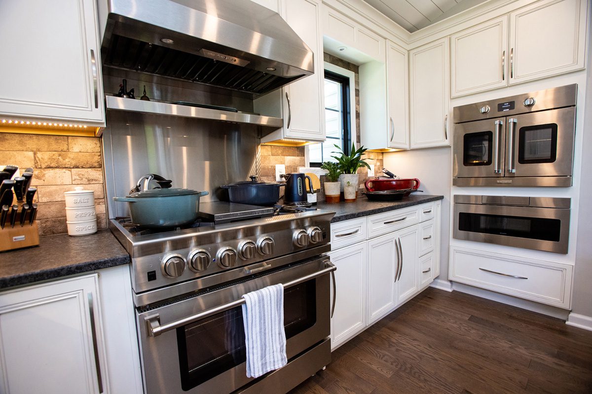 Modern kitchen with white cabinets, stainless steel double oven, large stove with pots, countertop appliances, and a window above the sink.