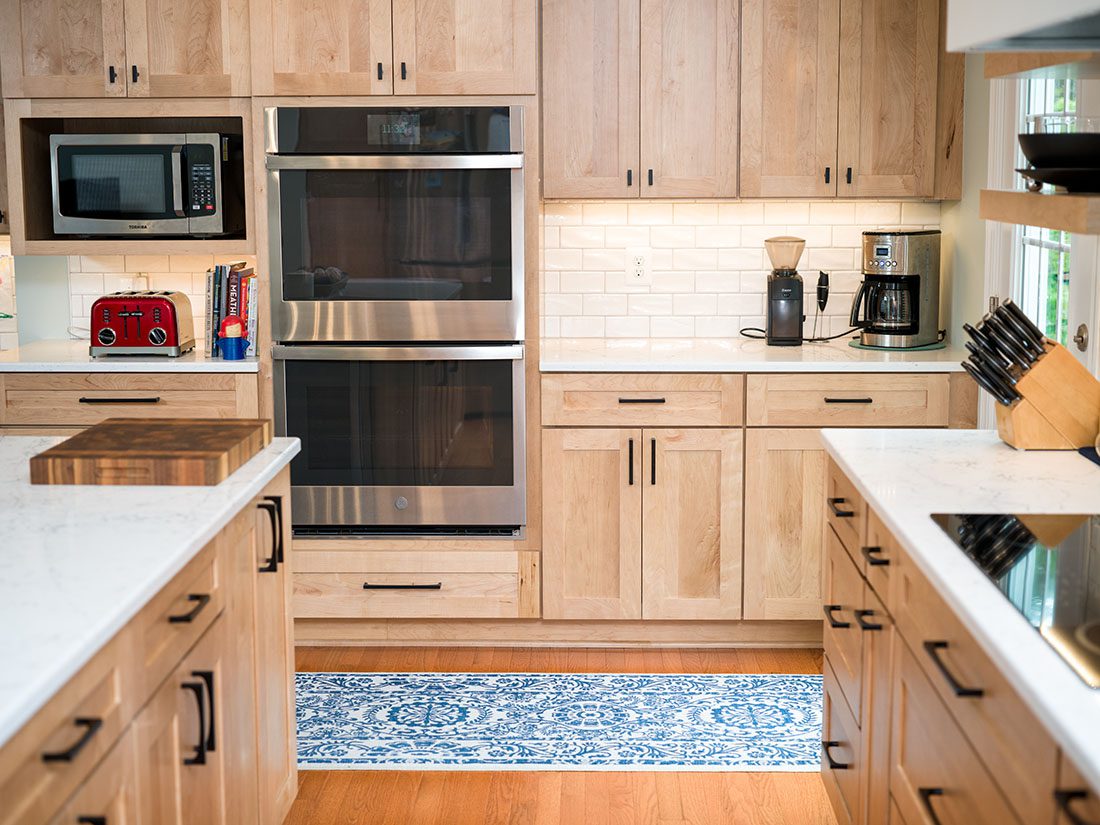 Modern kitchen with light wood cabinets, double wall oven, microwave, coffee maker, and appliances on marble countertops; blue patterned rug on wooden floor.