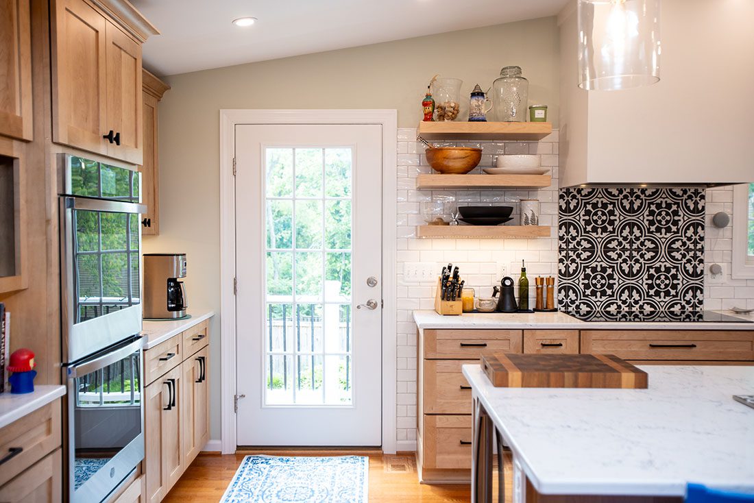 Modern kitchen with light wood cabinets, marble countertops, a patterned tile backsplash, open shelves with dishes, double oven, and a glass door letting in natural light.