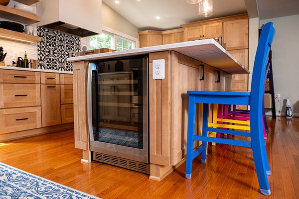 A modern kitchen with light wood cabinets, a kitchen island featuring a built-in wine fridge, and colorful chairs on a polished wooden floor.
