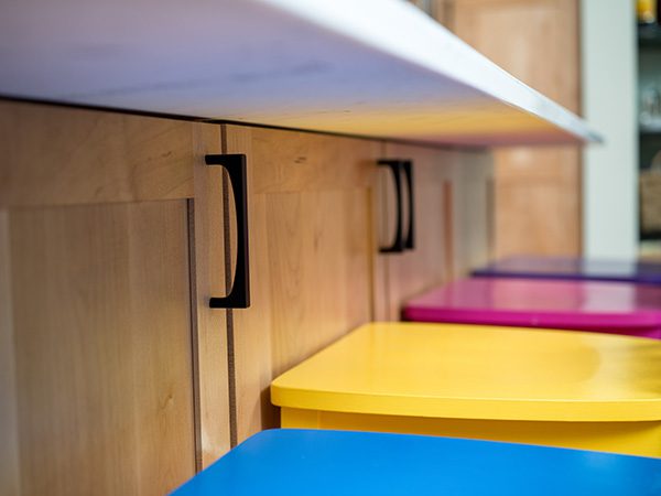 Close-up of a row of colorful stools—blue, yellow, pink, and purple—lined up under a white counter with wooden cabinets and black handles.