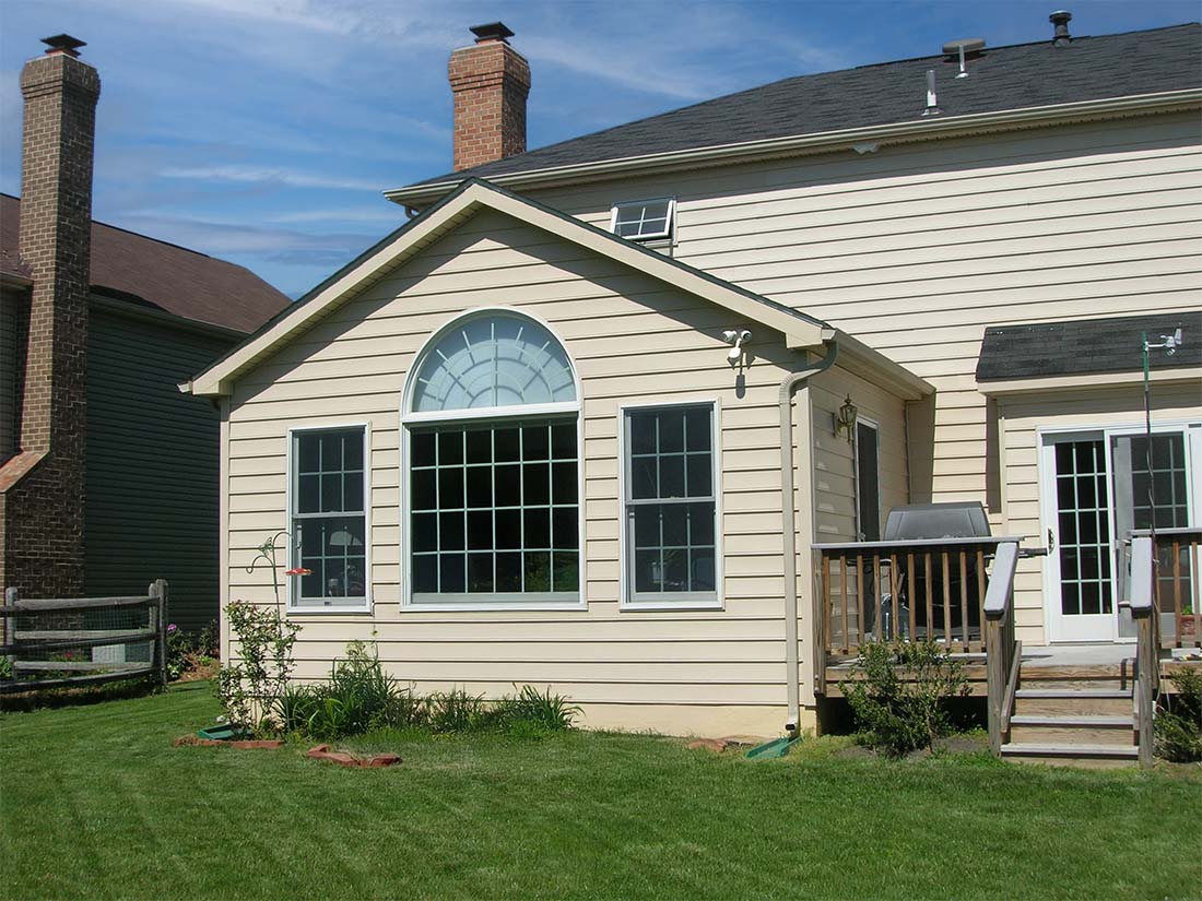 A beige house with a sunroom featuring a large arched window, a small wooden deck, and a grassy backyard—perfect for those considering elegant home additions in Fairfax VA.