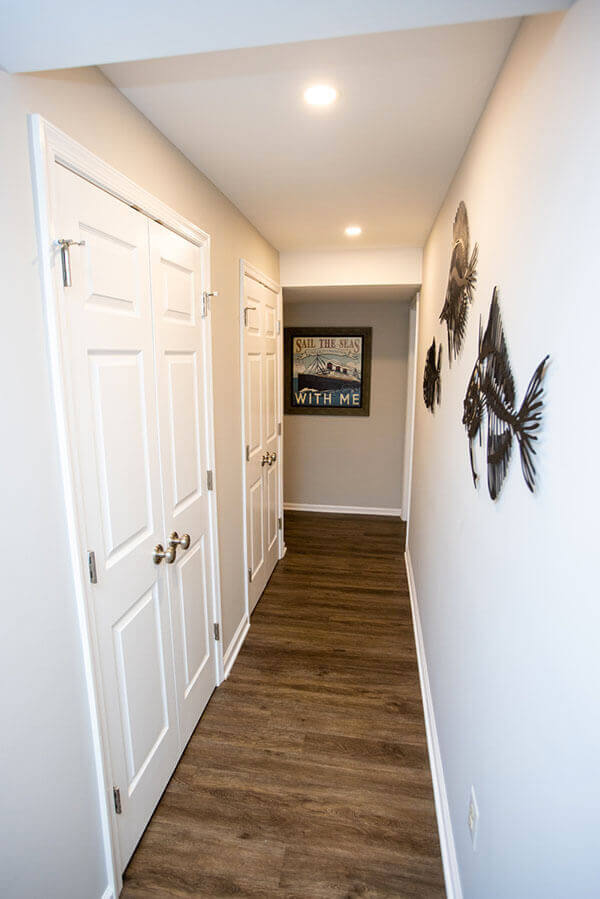 A narrow hallway with wood flooring, white doors on the left, decorative metal fish art on the right wall, and a framed nautical-themed sign at the end.