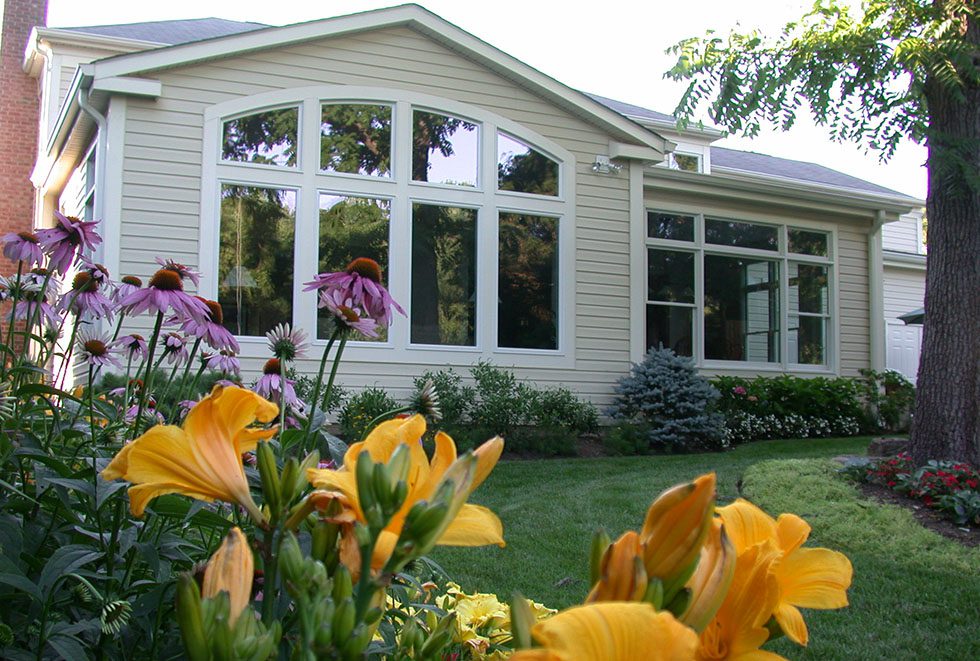 Large house with cream siding and many windows, viewed from a garden blooming with yellow lilies and purple coneflowers—perfect inspiration for home additions Fairfax VA homeowners desire.