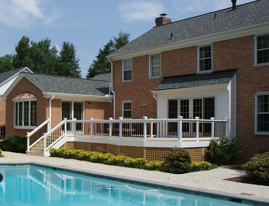 A brick house with white trim features a raised wooden deck with stairs, overlooking a rectangular backyard swimming pool and landscaped garden—perfect inspiration for stylish home additions Fairfax VA homeowners desire.