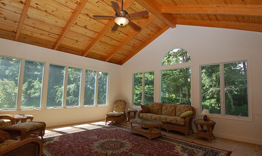 A sunroom with large windows, wicker furniture, a patterned rug, and a ceiling fan showcases beautiful trees outside—a perfect example of stylish home additions Fairfax VA residents love.