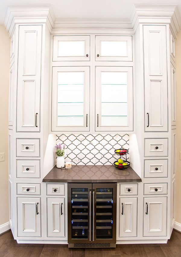 White kitchen cabinets with black countertops, a built-in wine fridge, glass-front uppers, and a patterned tile backsplash—perfect inspiration for kitchen remodeling McLean VA. Fruit and a plant add a fresh touch to the counter.