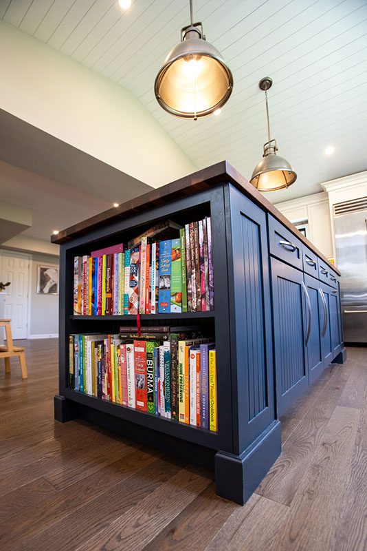 A navy blue kitchen island with built-in shelves containing colorful books, on a wooden floor under two pendant lights.