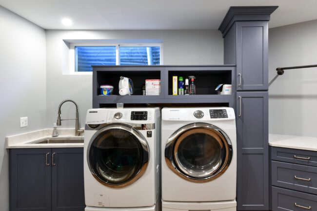 Modern laundry room with front-loading washer and dryer, blue cabinets, a sink with a gooseneck faucet, and shelves holding cleaning supplies under a small window.