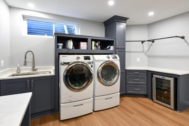 Modern laundry room with front-loading washer and dryer, a sink, shelving with cleaning supplies, countertop space, and a mini fridge on wood flooring.