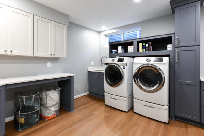 Modern laundry room with front-loading washer and dryer, cabinets, countertop, open shelves with supplies, and sorted laundry bags on a wood floor.