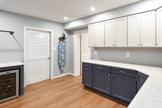 A modern laundry room with white and navy cabinets, marble countertops, wood flooring, an ironing board, and a small built-in wine fridge.