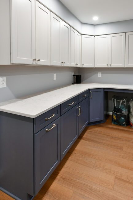 Modern kitchen with upper white cabinets, lower blue cabinets, white countertops, wood flooring, and reusable shopping bags stored in the corner.