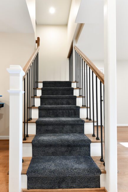 A straight staircase with dark carpet runner, white risers, wooden handrails, and black vertical balusters, leading up to the second floor of a house.
