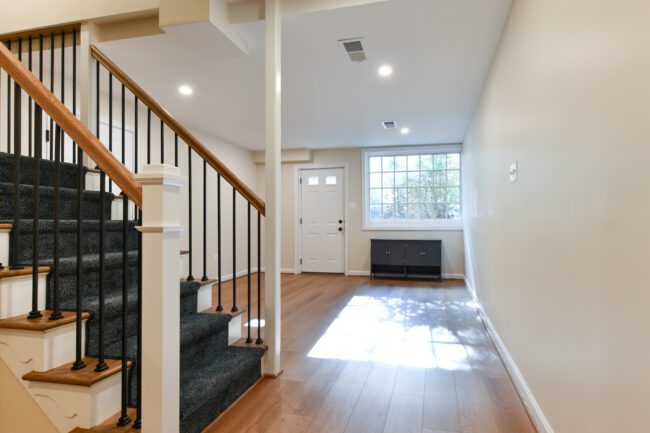 A modern entryway with wood flooring, a carpeted staircase with black railings on the left, a large window, and a white front door.
