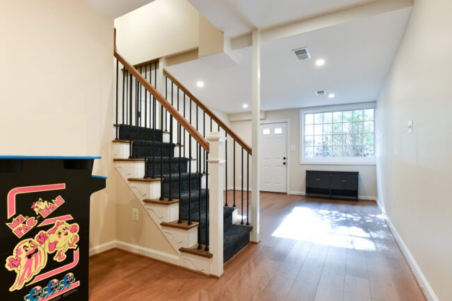 A well-lit entryway features a staircase with dark carpet, hardwood floors, a black cabinet, and a Ms. Pac-Man arcade machine in the foreground.