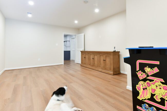 A spacious room with wood flooring, a wooden sideboard, an open door leading to another area, a black and white dog on the floor, and a Pac-Man arcade game in the corner.