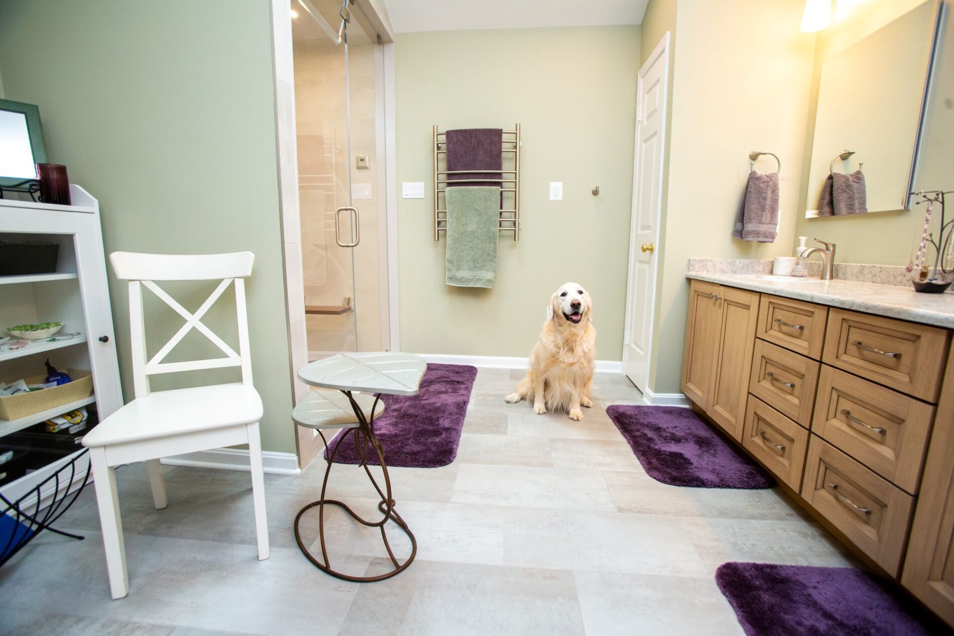 A golden retriever sits on the floor of a spacious bathroom with green walls, wooden cabinets, purple rugs, and modern fixtures—showcasing the charm that thoughtful remodeling vs building from scratch can bring to your space.