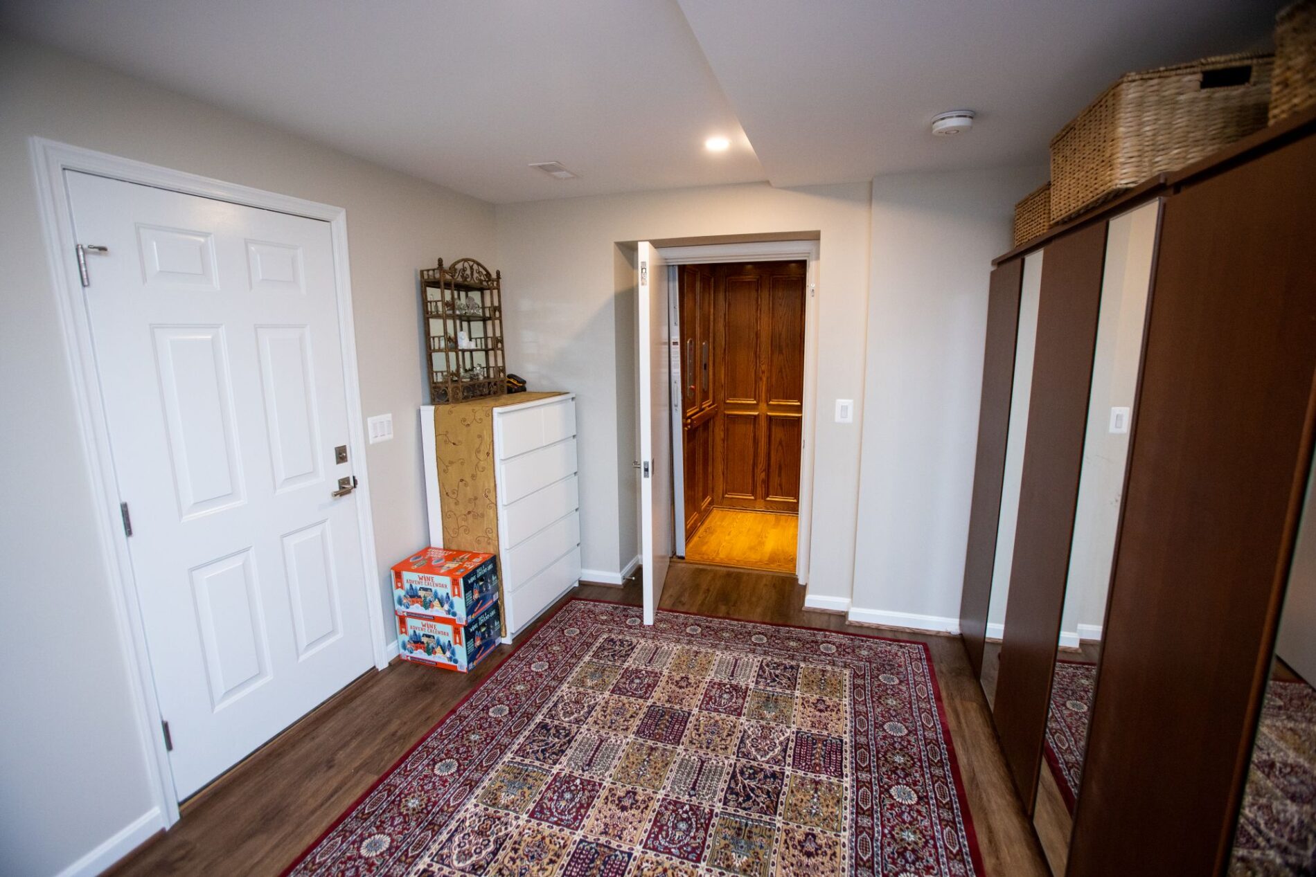 A room with a patterned rug, wardrobe, dresser, and a door open to a wood-paneled residential elevator—perfect for those considering remodeling vs building. Baskets top the wardrobe and boxes sit near the dresser for added storage.