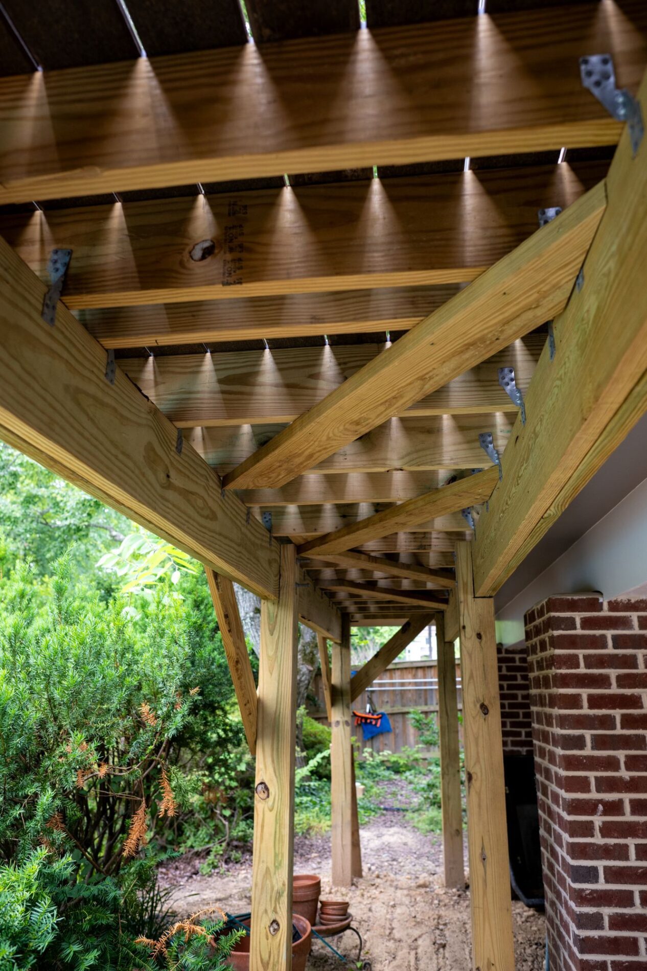 View of an unfinished wooden deck frame with exposed beams and support posts, perfect for anyone considering remodeling vs building new, situated next to a brick wall and overlooking a garden area.