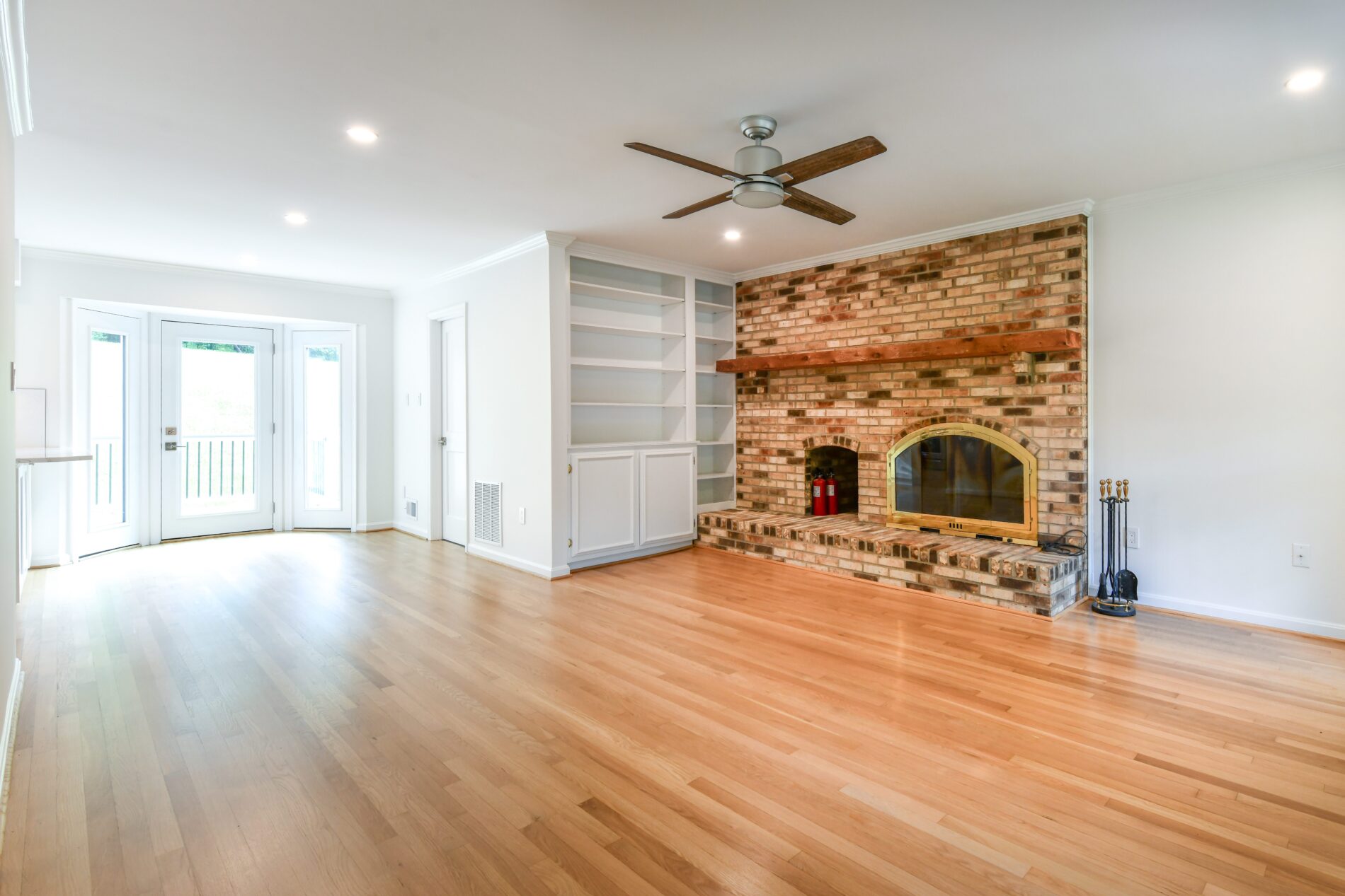 Spacious living room with light wood flooring, a brick fireplace, built-in white shelves, ceiling fan, and double glass doors leading outside.