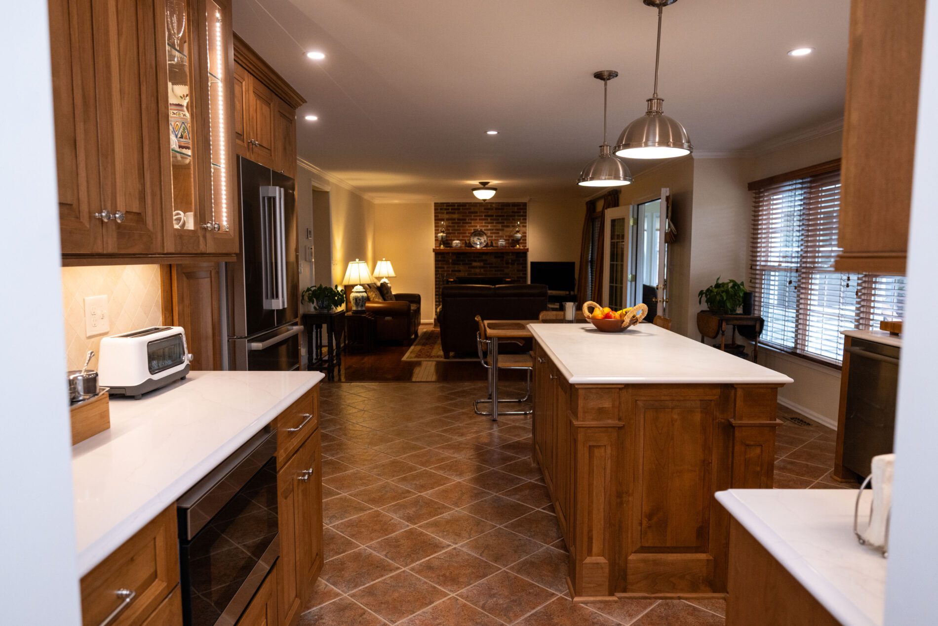 A spacious kitchen with wooden cabinets, white countertops, island, stainless steel appliances, and a view into a living area with a brick fireplace.