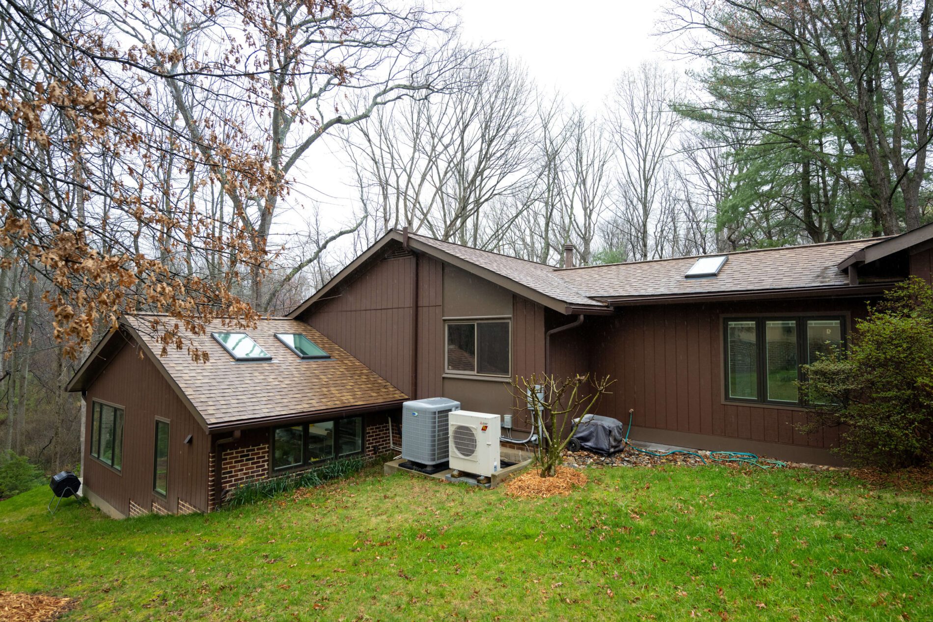 A brown house with a sloped roof, skylights, and large windows sits on a grassy yard, with HVAC units and trees in the background.