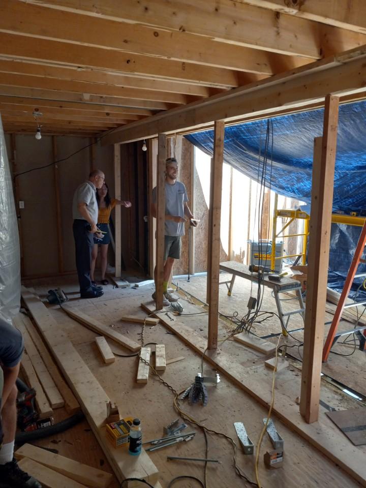 Three people stand inside a partially constructed building with exposed wooden beams and scattered tools, possibly discussing where to stay during a renovation amid the construction materials.