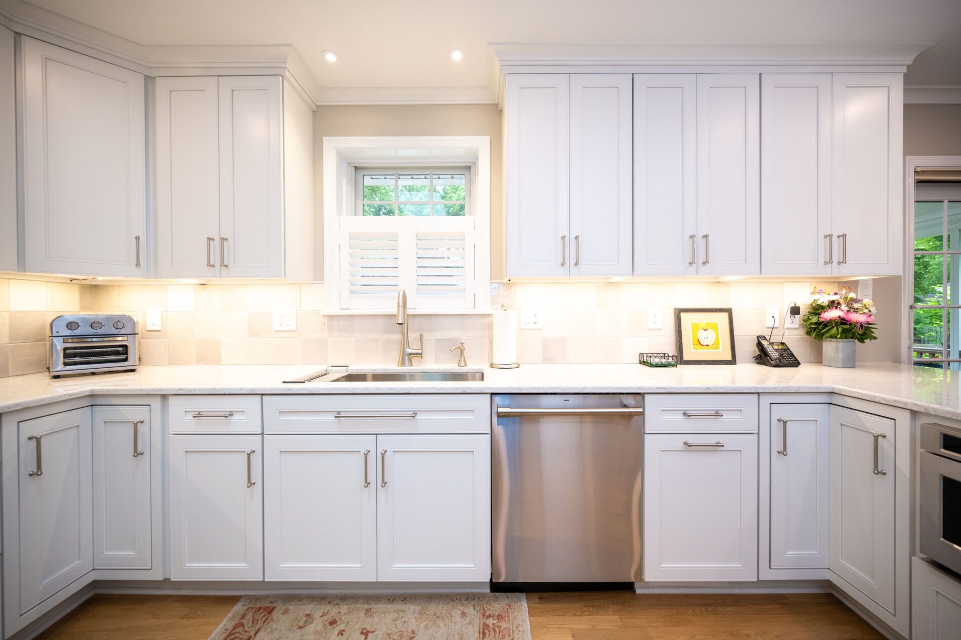 Modern kitchen with white cabinets, stainless steel appliances, a sink under a window, and light wood flooring. A toaster, framed art, and flowers sit on the countertop.