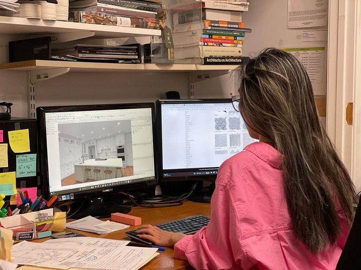 A person in a pink shirt sits at a desk working on two computer monitors, one displaying a 3D kitchen design and the other showing pattern options.