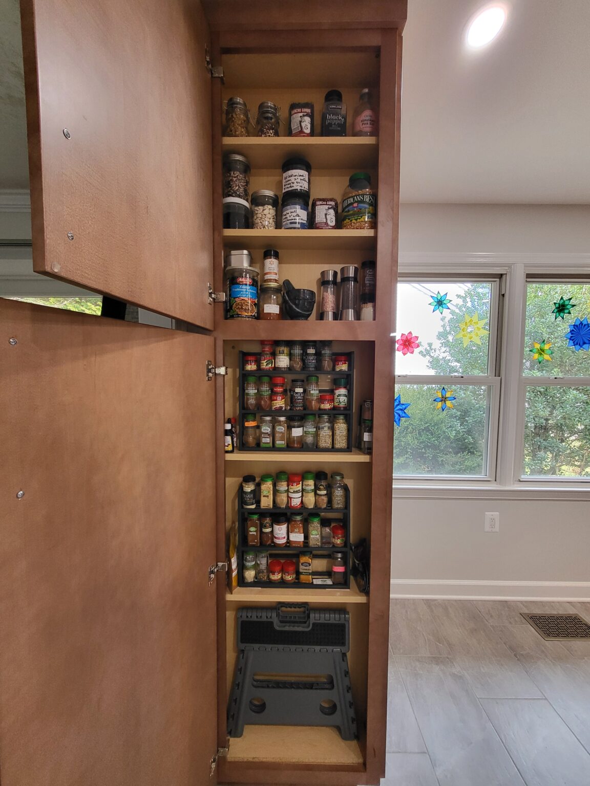 Tall kitchen cabinet with multiple shelves organized with spice jars and containers, and a folded step stool stored at the bottom.